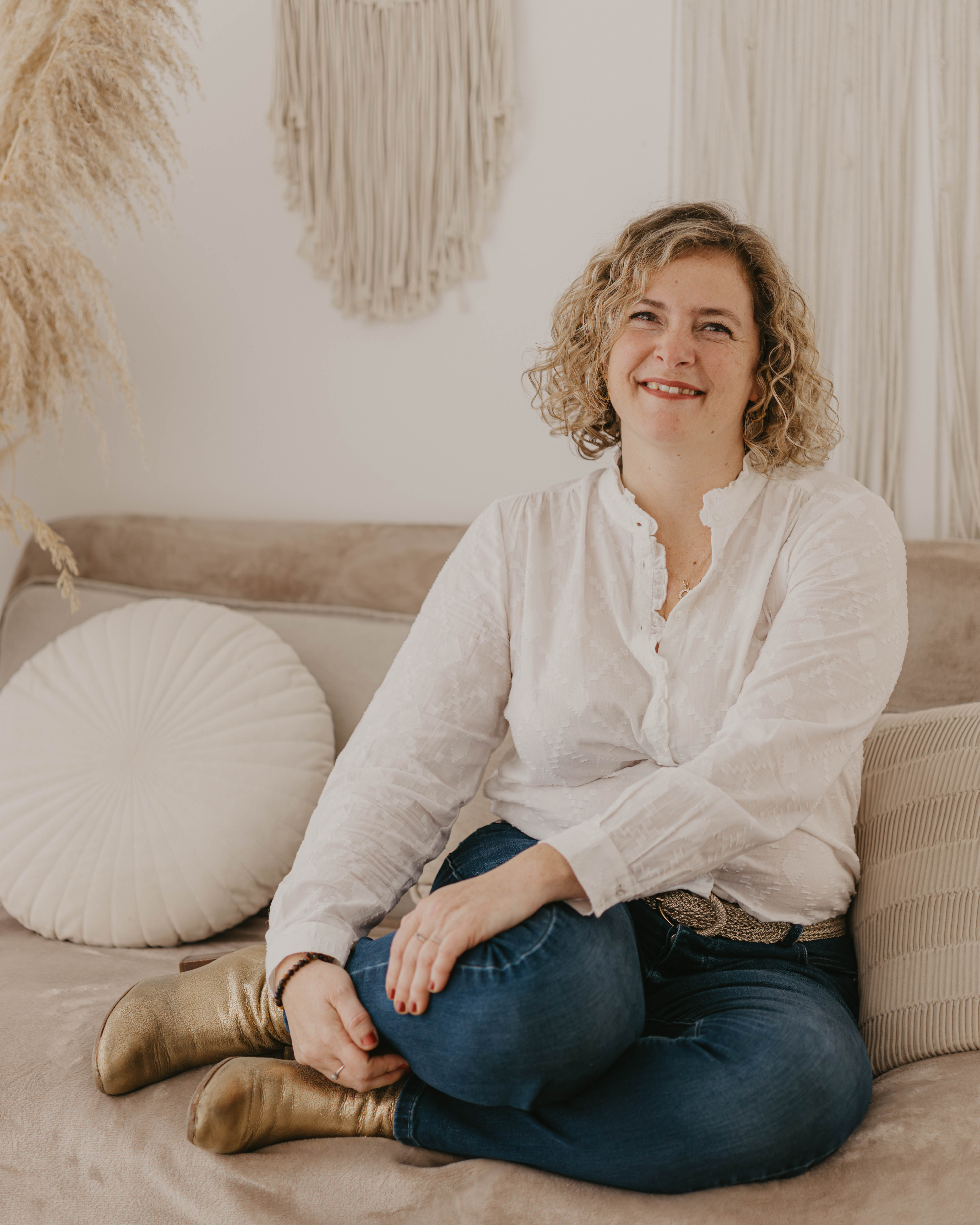 Woman sitting on a beige sofa, wearing a white blouse and jeans, with gold boots. Decorative wall hangings and cushions in the background.