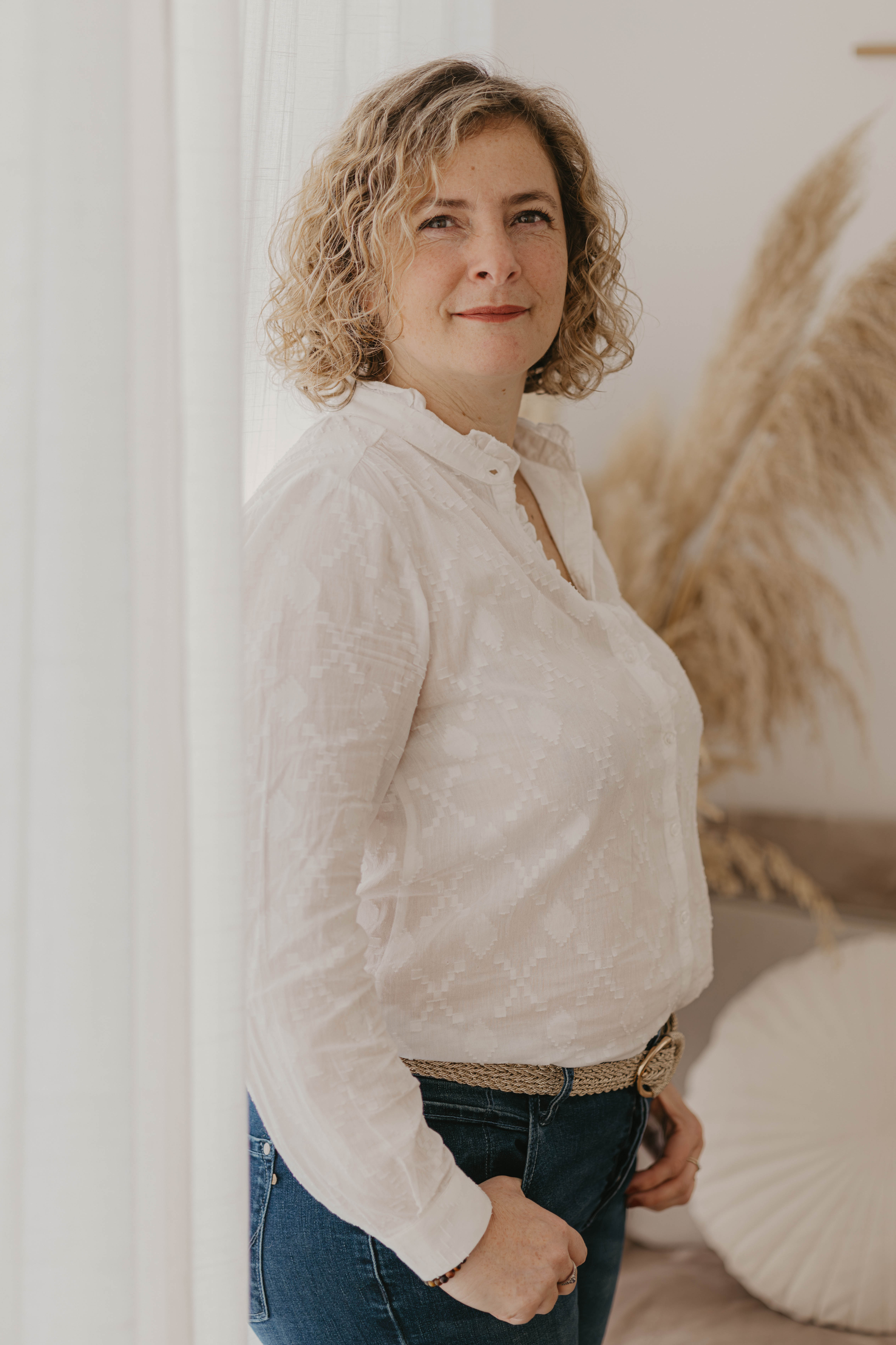 Woman with curly hair and a white blouse stands by a white curtain, smiling softly, with pampas grass in the background.